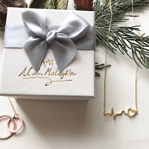 K&ouml;nnte beinhalten: Zwei goldene Halsketten in Geschenkboxen. Eine Halskette zeigt ein Herz mit einer Herzschlaglinie, die andere hat zwei ineinandergreifende Kreise. Die Boxen sind wei&szlig; mit einer silbernen Schleife und dem Text "N.d. McLaughlin" in Kursivschrift.