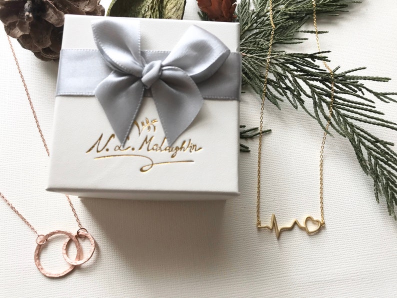 K&ouml;nnte beinhalten: Zwei goldene Halsketten in Geschenkboxen. Eine Halskette zeigt ein Herz mit einer Herzschlaglinie, die andere hat zwei ineinandergreifende Kreise. Die Boxen sind wei&szlig; mit einer silbernen Schleife und dem Text "N.d. McLaughlin" in Kursivschrift.