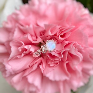 May include: A silver ring featuring a white opal gemstone set in a delicate floral design. The ring is resting on a pink carnation.