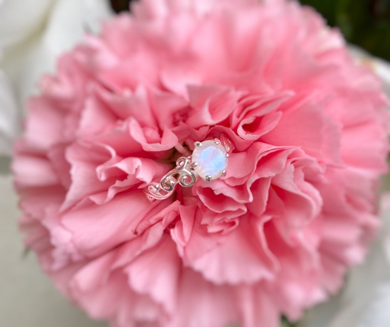 May include: A silver ring featuring a white opal gemstone set in a delicate floral design. The ring is resting on a pink carnation.