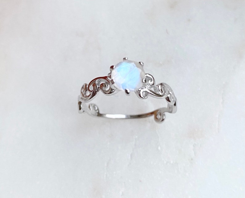 May include: A silver ring featuring a delicate, scroll-like design and a round, iridescent moonstone.