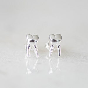 Könnte beinhalten: Ein Paar silberne Ohrstecker in Zahnform. Die Ohrringe sind klein und zart und wären ein tolles Geschenk für einen Zahnarzt oder jemanden, der Zähne liebt.