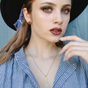 K&ouml;nnte beinhalten: Eine Frau tr&auml;gt ein blau-wei&szlig; gestreiftes Hemd mit einer goldenen Halskette mit einem kleinen Fl&uuml;gel-Anh&auml;nger. Sie tr&auml;gt einen schwarzen Hut und hat braunes Haar und Make-up.