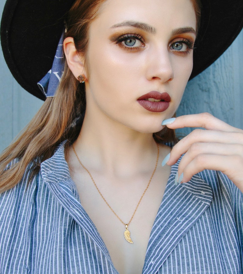 K&ouml;nnte beinhalten: Eine Frau tr&auml;gt ein blau-wei&szlig; gestreiftes Hemd mit einer goldenen Halskette mit einem kleinen Fl&uuml;gel-Anh&auml;nger. Sie tr&auml;gt einen schwarzen Hut und hat braunes Haar und Make-up.
