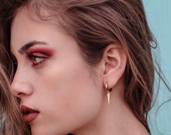 May include: A woman with long brown hair wearing gold hoop earrings with a pointed pendant. The earrings are a simple design with a small hoop and a long, thin pendant that ends in a sharp point. The woman is wearing red eyeshadow and has a neutral expression.