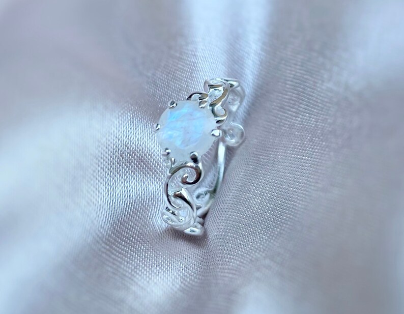 May include: A silver ring with a delicate, ornate design features a round, iridescent moonstone.