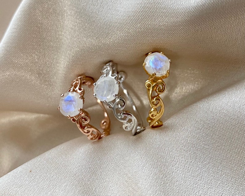 May include: Three rings featuring a moonstone gemstone set in a delicate, ornate, scroll-like design. The rings are in gold, silver, and rose gold.