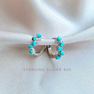 May include: A pair of sterling silver hoop earrings with turquoise stones. The earrings are on a white fabric background. STERLING SILVER 925 is written below the earrings.