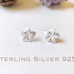 Puede incluir: Un par de pendientes de botón de plata de ley con forma de flor. Cada pendiente presenta un diseño de flor de cinco pétalos. El texto "STERLING SILVER 925" está impreso debajo.