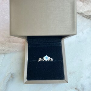 May include: A silver ring featuring a white moonstone set in a delicate floral design. The ring is presented in a black velvet ring box.