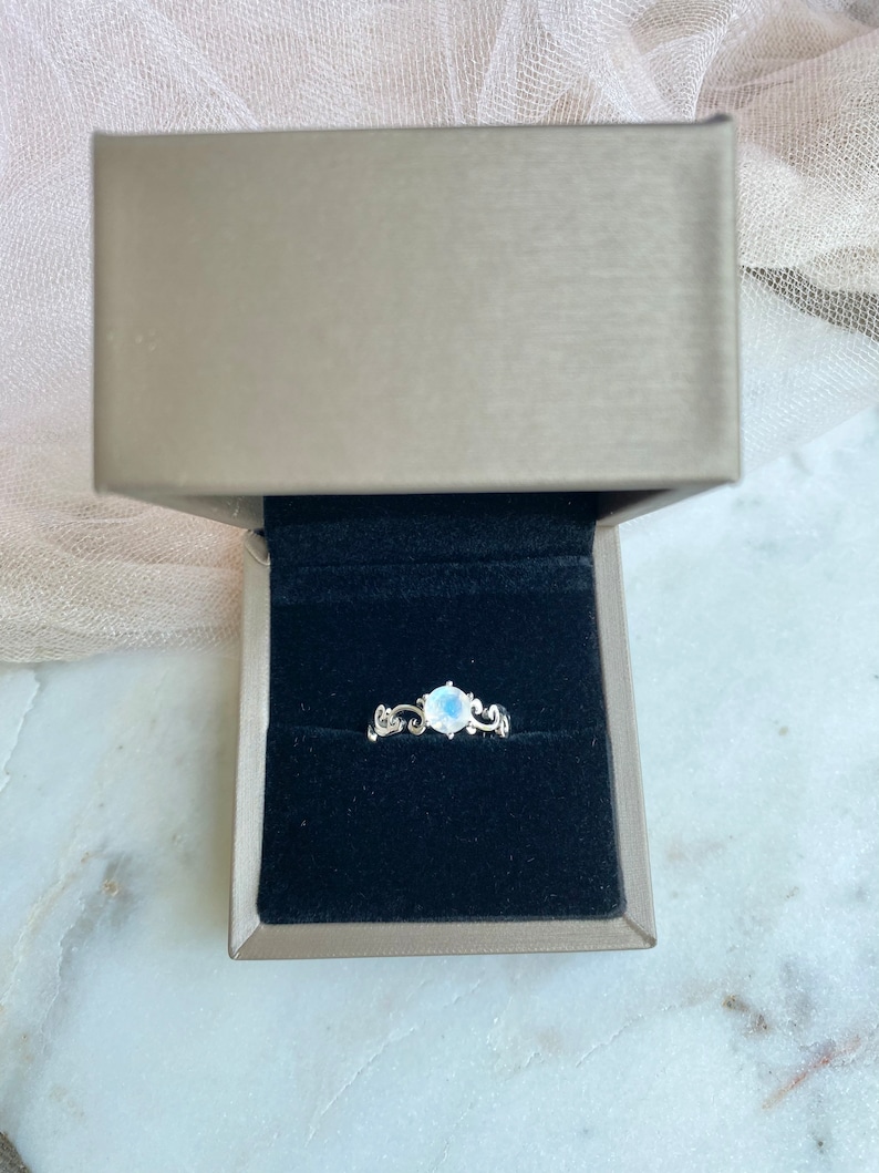 May include: A silver ring featuring a white moonstone set in a delicate floral design. The ring is presented in a black velvet ring box.