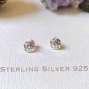 May include: A pair of sterling silver 925 stud earrings with a woven knot design. The text "STERLING SILVER 925" is printed in brown below the earrings. The background is a white textured surface.