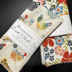 May include: A handmade dish cloth set made of 100% cotton, featuring a cream background with colorful chicken and floral patterns. A white paper band wraps around the cloths with the text "HANDMADE Dish Cloth Set 100% COTTON MACHINE WASH & DRY".