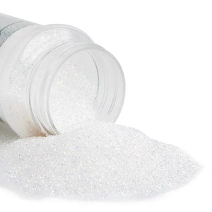 May include: A clear plastic container tipped over, spilling iridescent white glitter onto a white surface. The glitter sparkles with hints of blue and silver, creating a shimmering effect. The container is partially visible, showing its open top.