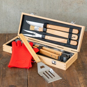 May include: A wooden barbecue tool set with stainless steel utensils and wooden handles. The set includes a spatula, fork, brush, and knife, along with a red silicone oven mitt. The tools are stored in a wooden box.