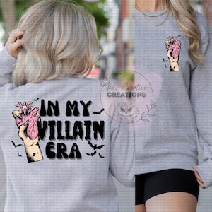 May include: A gray sweatshirt with the text "In My Villain Era" and a graphic of a hand holding a heart. The graphic is in black, pink, and white.