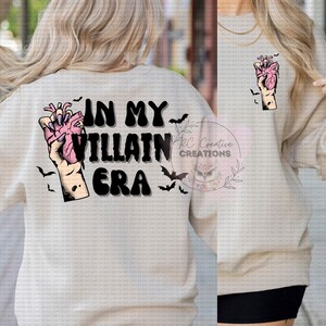 May include: A white sweatshirt with the text "In My Villain Era" in black letters. A black and pink hand holding a heart is on the left side of the text. There are black bats around the text.