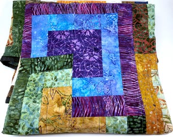 May include: A quilt made of colorful fabric squares in shades of purple, blue, green, and brown. The quilt is made in a log cabin pattern with a square in the center.