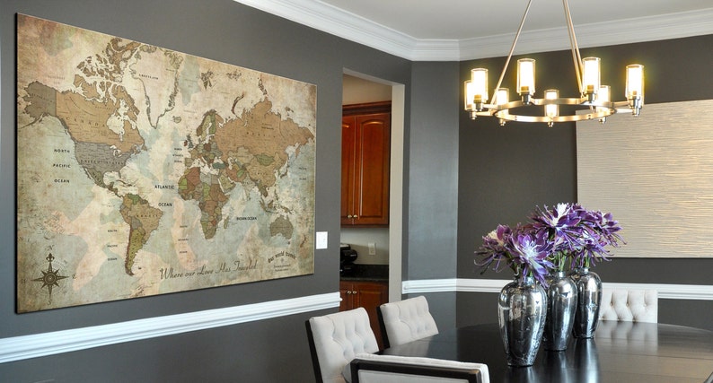 May include: A large world map print with a vintage style design hangs on a gray wall in a dining room. The map is printed on a textured paper with a brown and beige color scheme. The map is surrounded by a white trim and the text "Where our love has traveled" is printed at the bottom of the map.