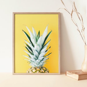 Könnte beinhalten: Gerahmter Kunstdruck mit einer Ananas vor einem leuchtend gelben Hintergrund. Die Ananas hat grüne und weiße Blätter und eine strukturierte, gelbbraune Basis. Der Rahmen ist helles Holz. Eine Vase mit getrocknetem Gras und ein Buch sind im Hintergrund.