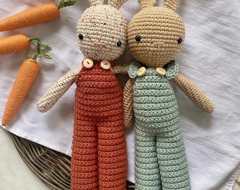 May include: Two crocheted bunny plushies wearing overalls. One bunny is beige with brown eyes and a light blue overall. The other bunny is white with brown eyes and a red overall.