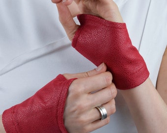 May include: A pair of fingerless gloves in a vibrant red colour with a textured, pebbled surface. These short gloves cover the palm and back of the hand, leaving the fingers and thumb exposed, and feature stitched edges along the openings.