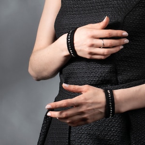 Puede incluir: Brazaletes negros con filas de pequeños adornos brillantes. Los brazaletes están hechos de un material suave y oscuro y se usan en ambas muñecas. El fondo es gris liso.