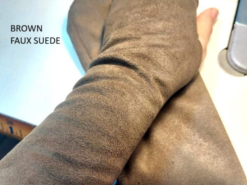 May include: A pair of brown faux suede fingerless gloves with a soft, matte texture. The fabric is flexible and covers the forearm, featuring a thumb opening. The text BROWN FAUX SUEDE is printed in black on the upper left side of the image.