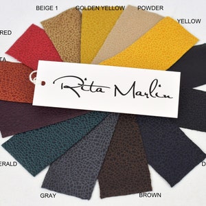 May include: A color swatch sample set featuring twelve textured leather-like material strips arranged in a fan shape. The colors include red, beige, golden yellow, powder, yellow, black, dark blue, brown, gray, emerald, and burgundy. A white tag with the text Rita Marlin is attached.