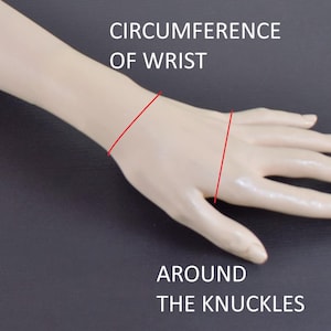 May include: A beige mannequin hand and forearm showing measurement lines. Text reads CIRCUMFERENCE OF WRIST above a red line around the wrist, and AROUND THE KNUCKLES below a red line across the hand, indicating where to measure for jewelry sizing.