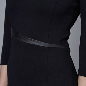 May include: A black dress featuring three-quarter length sleeves and a fitted bodice. The garment includes a distinct, angled satin-finish waistband detail that wraps across the midsection, providing a subtle contrast to the matte fabric.