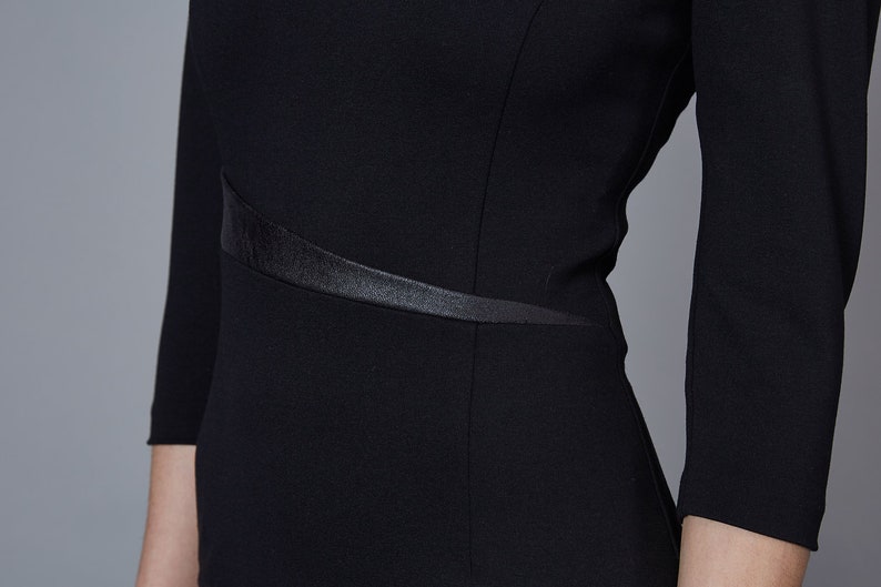 May include: A black dress featuring three-quarter length sleeves and a fitted bodice. The garment includes a distinct, angled satin-finish waistband detail that wraps across the midsection, providing a subtle contrast to the matte fabric.