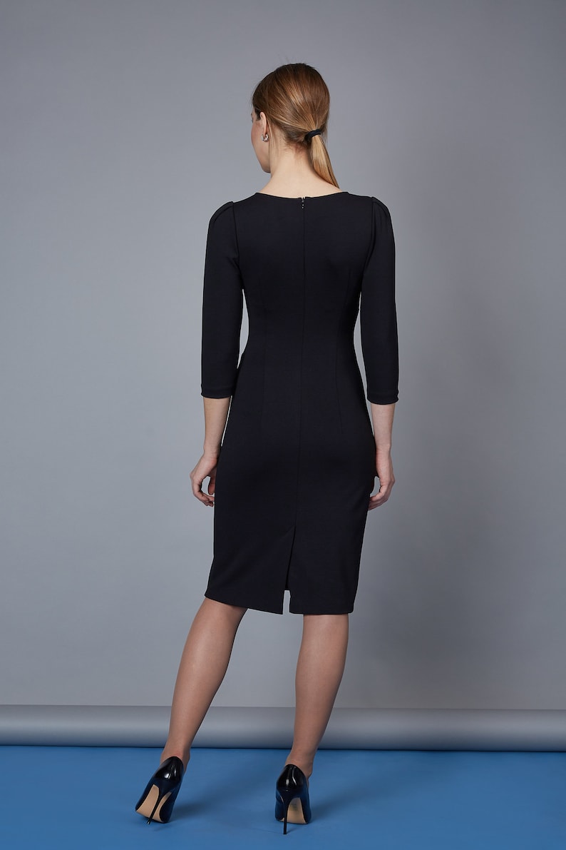 May include: A black knee-length dress with three-quarter length sleeves, a round neckline, and a vertical zipper closure along the center back. The garment features a straight silhouette with a small slit at the bottom hem of the back panel.
