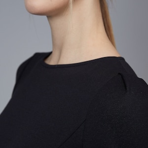 May include: A solid black top featuring a simple round neckline and subtle pleating detail at the shoulder seams. The garment is constructed from a smooth, opaque fabric that provides a clean, structured look across the chest and shoulder area.
