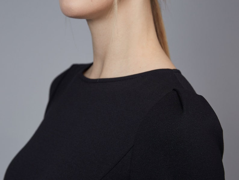 May include: A solid black top featuring a simple round neckline and subtle pleating detail at the shoulder seams. The garment is constructed from a smooth, opaque fabric that provides a clean, structured look across the chest and shoulder area.