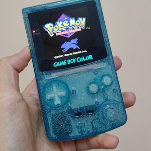 Op de afbeelding: Een doorschijnende, blauwgroene Game Boy Color handheld spelcomputer met een glitterafwerking. Het scherm toont het titelscherm van Pokémon Crystal Version met de tekst 2001 GAME FREAK inc. Het apparaat heeft een richtingspad en twee knoppen.