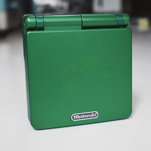 May include: A green Nintendo Game Boy Advance SP handheld game console. The console is closed and has a glossy finish. A logo is visible on the front, and the Nintendo logo is at the bottom.
