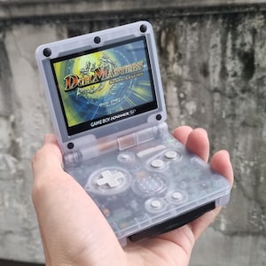 May include: A clear Game Boy Advance SP handheld console, showing a game screen with the text "Duel Masters". The console has a D-pad, buttons, and the text "GAME BOY ADVANCE SP".