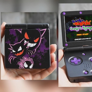 Può includere: Game Boy Advance SP nero con un design di Gengar viola. La console è aperta, mostrando il gioco "Banjo-Kazooie: Grunty's Revenge". Sono visibili pulsanti e dettagli viola.