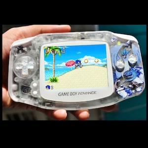 May include: A clear Game Boy Advance handheld console displaying a Sonic the Hedgehog game on its screen. The console has a white body with a clear front and a blue and white design on the right side. The game shows Sonic running on a beach.