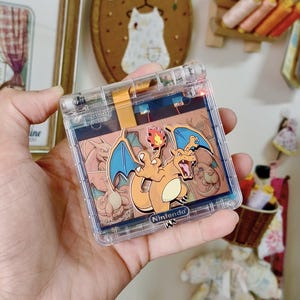 May include: A clear plastic handheld gaming console featuring a Charizard graphic on the screen. The device has a hinged design with a visible internal ribbon cable and a Nintendo logo printed on the lower edge of the screen frame.