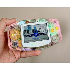 May include: A clear Game Boy Advance handheld console with a pink and yellow gradient shell. The screen displays "FINAL FANTASY I-II DAWN OF SOULS". The console has a D-pad, buttons, and a power switch.