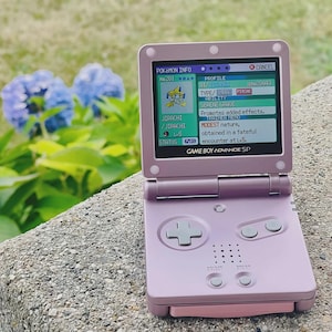May include: A lavender Game Boy Advance SP handheld console, open to display a Pokémon profile. The screen shows a Jirachi character with details like type, ability, and trainer memo. The console is on a stone surface with blue flowers in the background.