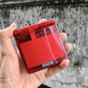 May include: A handheld video game console with a translucent red plastic shell. The device features a square, clamshell design with a hinge at the top. The word Nintendo is printed in white text on a small oval badge located on the lower front cover.