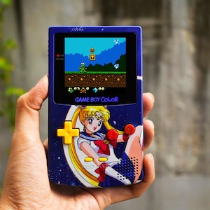 Könnte beinhalten: Eine violette Game Boy Color Handheld-Spielkonsole mit einer Sailor Moon-Grafik auf der Vorderseite. Das Gerät verfügt über ein gelbes Steuerkreuz, zwei rote Aktionstasten sowie gelbe Select- und Start-Tasten. Der Bildschirm zeigt ein Spiel.