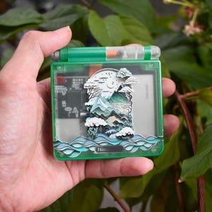 May include: A green handheld gaming device with a landscape diorama inside. The diorama features a mountain range, trees, and water, with a crescent moon in the sky. The device has the word "Nintendo" printed on the bottom.