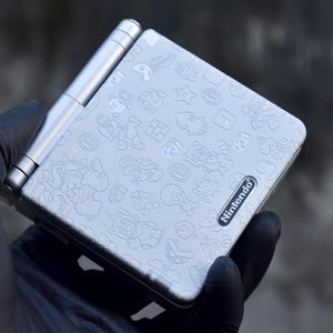 May include: Silver Nintendo Game Boy Advance SP handheld game console with a Mario-themed design. The console features a flip-up screen and a variety of embossed cartoon characters. The Nintendo logo is visible on the bottom right.