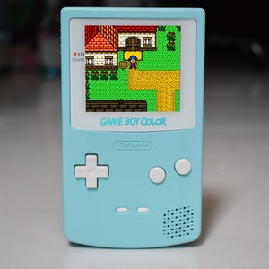 Puede incluir: Una consola de juegos portátil Game Boy Color azul claro con una placa frontal blanca. La pantalla muestra un videojuego pixelado con una casa y un personaje. La consola tiene un pad direccional en forma de cruz blanco, dos botones blancos y un altavoz.