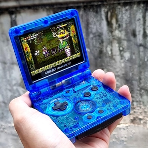 May include: A translucent blue Game Boy Advance SP handheld gaming console with a clear blue shell. The screen displays a video game with a green, yellow, and brown color scheme. The text "GAME BOY ADVANCE SP" is visible on the console.