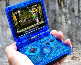 Könnte beinhalten: Eine durchsichtige blaue Game Boy Advance SP Handheld-Spielkonsole mit einem durchsichtigen blauen Gehäuse. Der Bildschirm zeigt ein Videospiel mit einem grünen, gelben und braunen Farbschema. Der Text "GAME BOY ADVANCE SP" ist auf der Konsole sichtbar.